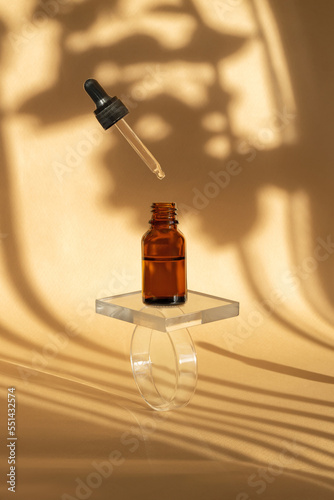 Serum in amber-colored glass bottle with levitating dropper lid on transparent prism stand. Essential oil for care of women's skin on beige background, in rays of sunlight, reflection. Vertical frame