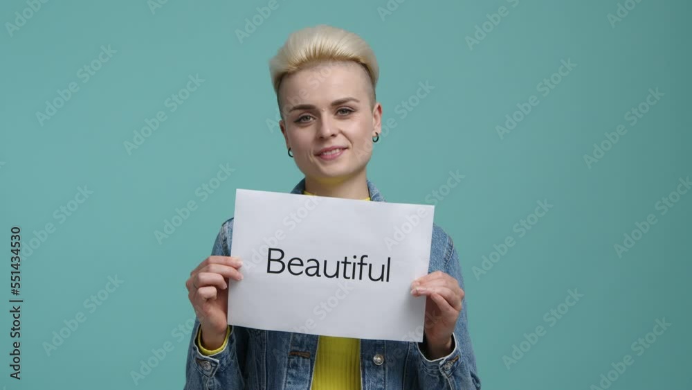 Close-up shot of caucasian, blonde-haired girl looking cheerful and smiling. Woman with positive facial expression holding a card with a Beautiful word. High quality 4k footage Close-up shot of caucasian, blonde-haired girl looking cheerful and smiling. Woman with positive facial expression holding a card with a Beautiful word. High quality 4k footage