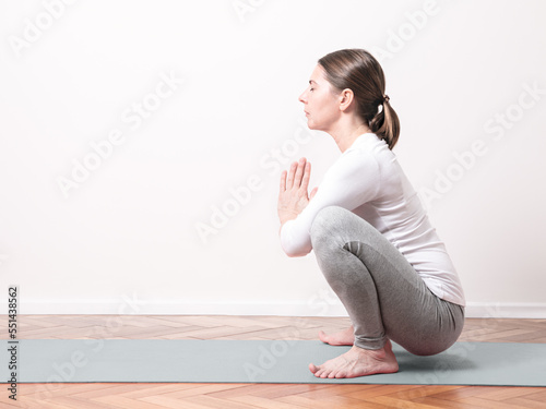 Young woman practices yoga in Malasana frog posture over gray mat in a empty room with wood floor photo