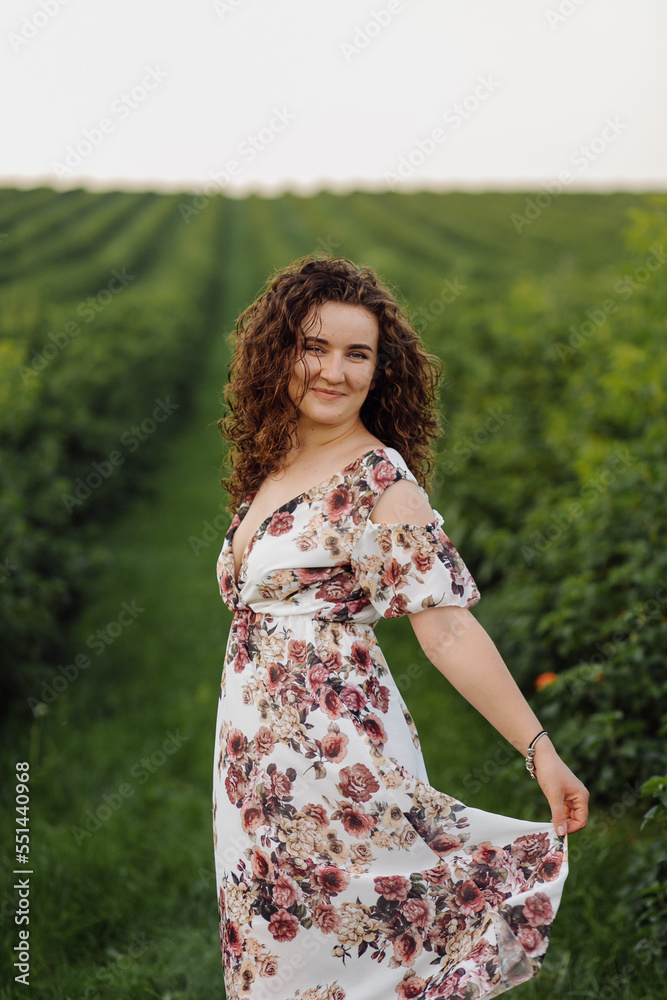 Happy young woman with brown curly hair, wearing a dress, posing outdoors in a garden Happy young woman with brown curly hair, wearing a dress, posing outdoors in a garden