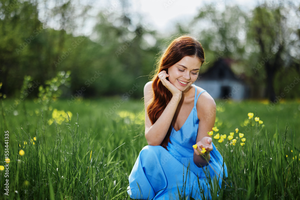 Woman in spring in nature smile with teeth sitting in a field on the green grass with flowers and smiling happy sunset in a blue dress with red hair Woman in spring in nature smile with teeth sitting in a field on the green grass with flowers and smiling happy sunset in a blue dress with red hair
