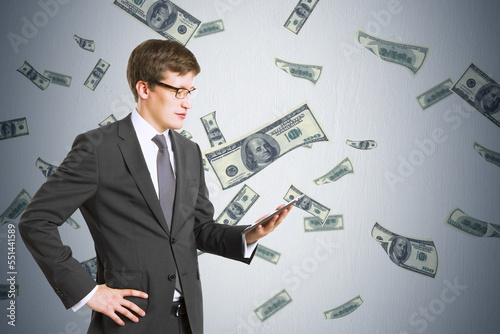 Attractive young european businessman using cellphone with creative dollar bill rain on concrete wall background. Millionaire, rich and cash concept.