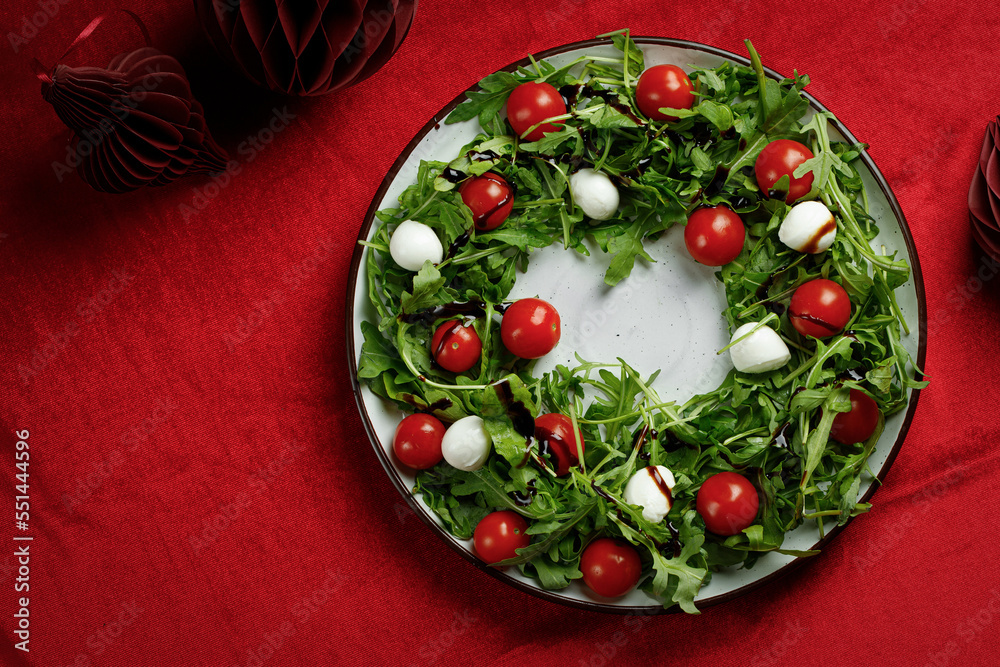Christmas wreath of Salad with arugula, cherry tomatoes and mozzarella cheese on red cloth table with red balls. Christmas food background. Christmas wreath of Salad with arugula, cherry tomatoes and mozzarella cheese on red cloth table with red balls. Christmas food background.