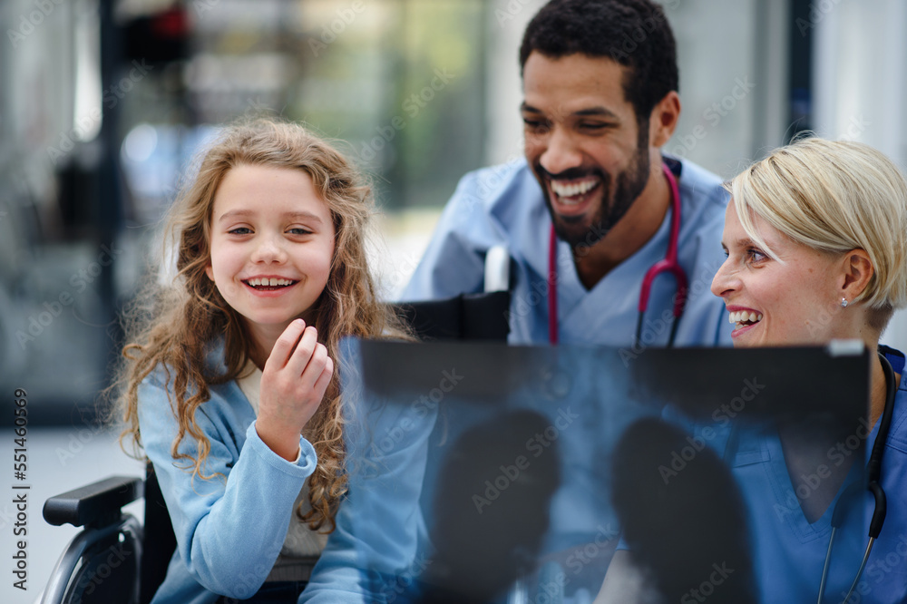 Young multiracial doctor with his colleague showing x-ray image of lungs to little girl on wheelchair.