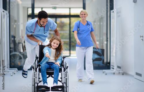 Young doctor pushing little girl at wheelchair at pediatric corridor, having fun.
