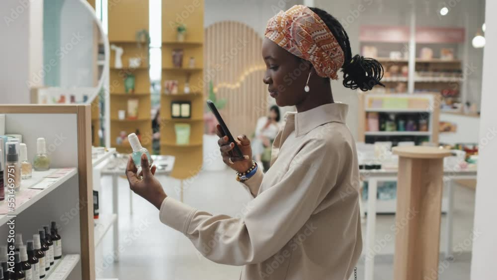 Side view of young black woman with smartphone scanning qr codes on cosmetic products while standing in front of large display with self care items in supermarket