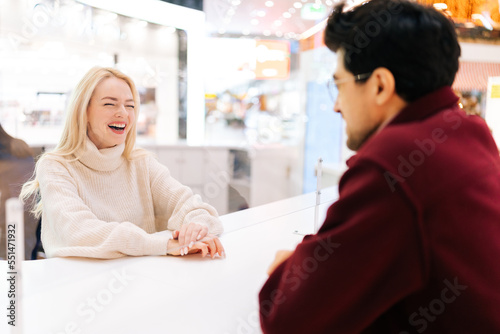 Back view from shoulder of unrecognizable young man chatting through glass partition with cheerful blonde woman standing in hall of shopping mall. Concept of lifestyle social distancing and work.