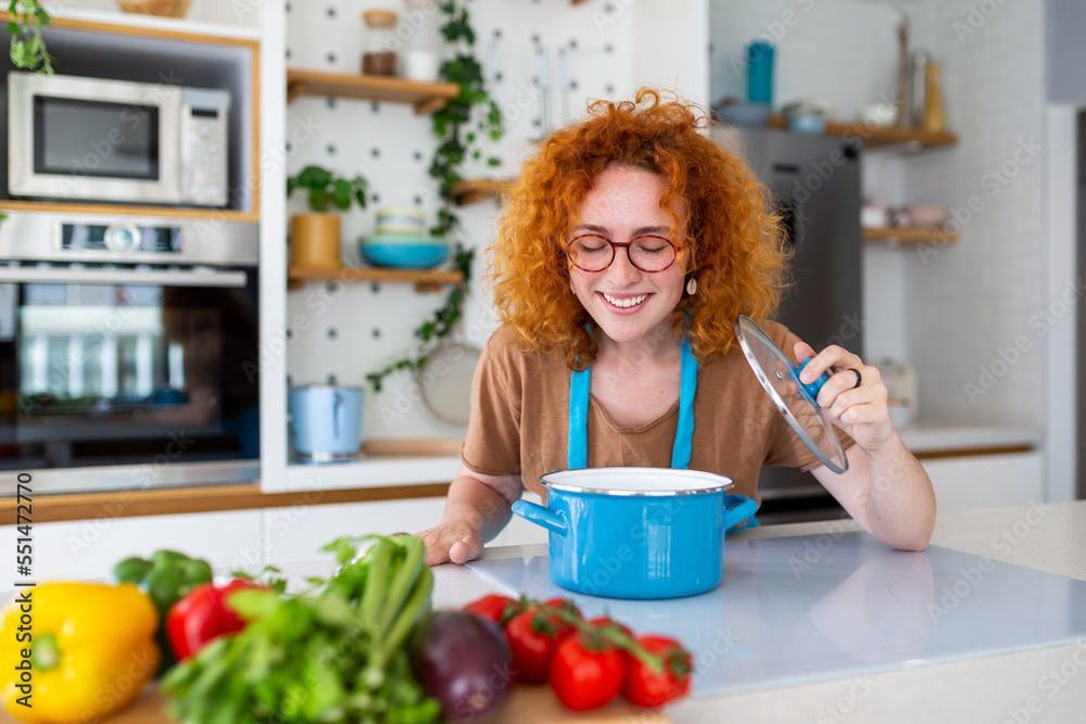 Cheerful young pretty female in apron prepare lunch and smell dish in kitchen at home. Woman cooking dinner for family at new recipe at home.