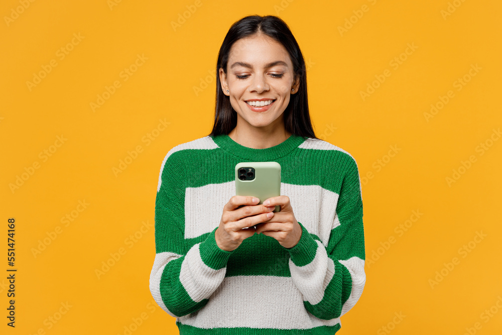 Young smiling brunette latin woman wear casual cozy green knitted sweater hold in hand use mobile cell phone in mint case isolated on plain yellow background studio portrait People lifestyle concept. Young smiling brunette latin woman wear casual cozy green knitted sweater hold in hand use mobile cell phone in mint case isolated on plain yellow background studio portrait People lifestyle concept.
