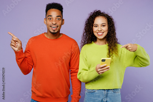Young couple two friends family man woman of African American ethnicity wear casual clothes together hold in hand use mobile cell phone point finger aside isolated on pastel plain purple background.