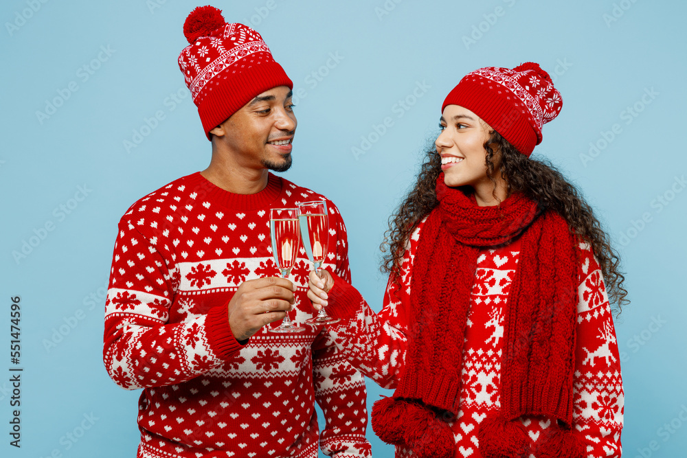 Merry young fun couple two man woman wear red Christmas sweater Santa hat posing drink champagne clink raise toast isolated on plain pastel light blue background. Happy New Year 2023 holiday concept.