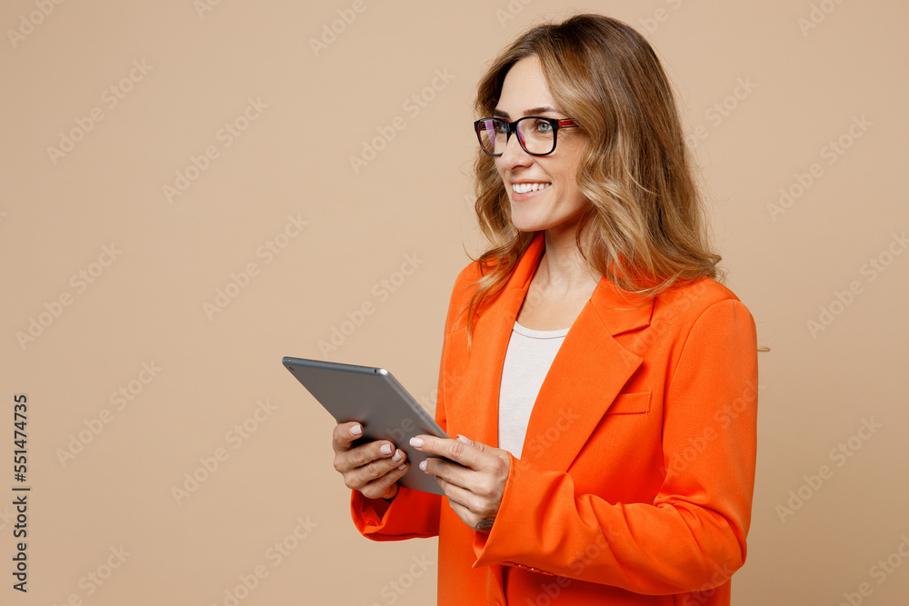 Side view young happy fun employee business woman corporate lawyer wear classic formal orange suit glasses work in office hold use digital tablet pc computer isolated on plain beige background studio. Side view young happy fun employee business woman corporate lawyer wear classic formal orange suit glasses work in office hold use digital tablet pc computer isolated on plain beige background studio.