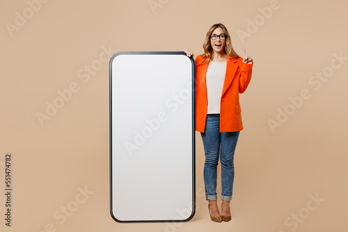 Full body fun young employee business woman corporate lawyer in formal orange suit work in office big huge blank screen mobile cell phone with area point finger up isolated on plain beige background