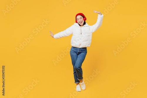 Full body cheerful young woman of Asain ethnicity wear white padded windbreaker jacket red hat headphones listen to music dance isolated on plain yellow background studio. People lifestyle concept.