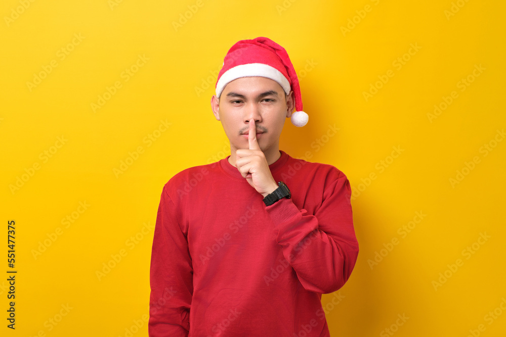 Young Asian man in Santa hat showing silence gesture, holding finger near lips over yellow studio background. celebration Christmas holiday and New Year concept Young Asian man in Santa hat showing silence gesture, holding finger near lips over yellow studio background. celebration Christmas holiday and New Year concept