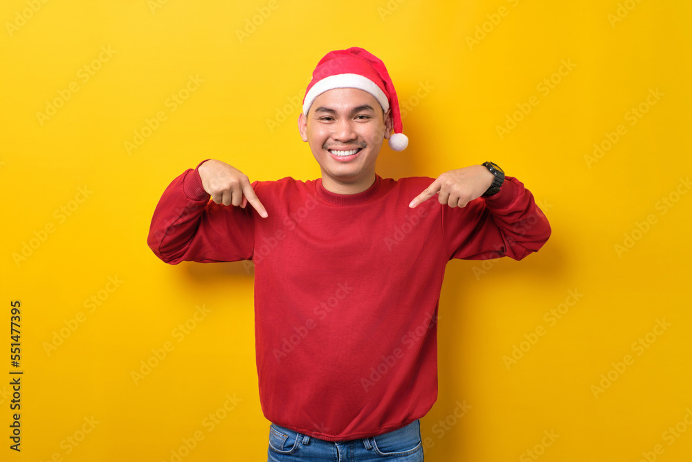 Cheerful young Asian man in Santa hat pointing fingers down and advertising something over yellow studio background. celebration Christmas holiday and New Year concept