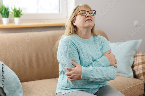 Indoor portrait of happy elderly woman hugging herself, giving support, showing self-respect and love, sitting on couch with closed eyes in urquoise sweater. Human emotions, body language photo