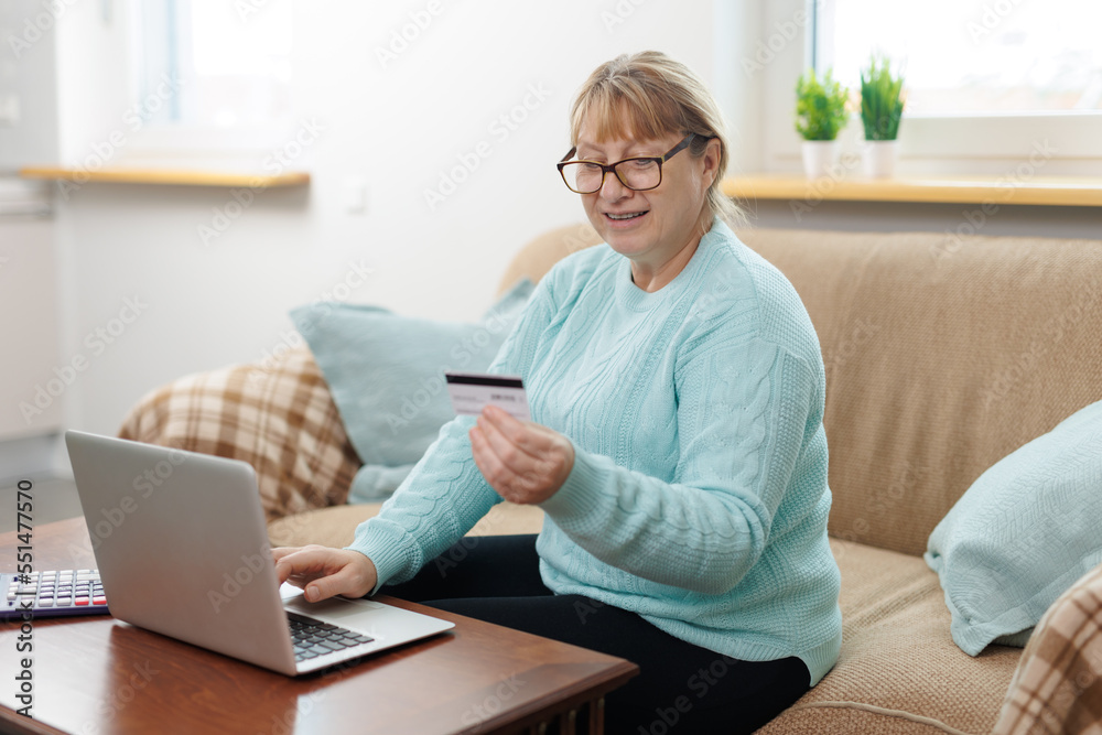 Clever senior satisfied woman sitting in the bright room on the sofa using the laptop and holding credit card. Clever senior satisfied woman sitting in the bright room on the sofa using the laptop and holding credit card.