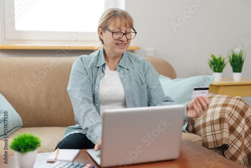 Clever senior satisfied woman sitting in the bright room on the sofa using the laptop and holding credit card.