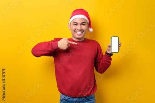Cheerful young Asian man in Santa hat pointing fingers at smartphone with blank screen over yellow studio background. celebration Christmas holiday and New Year concept © Sewupari Studio