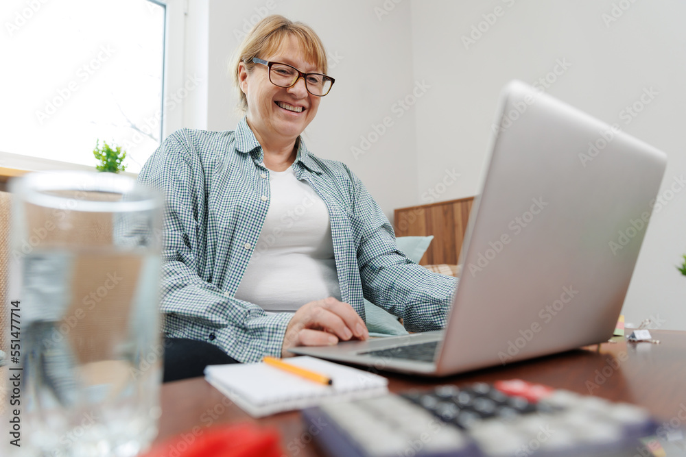 Happy mature businesswoman accountant in glasses and turquoise jumper sitting on the couch with laptop, taking notes, holding papers, contract studying or working remotely online from home Happy mature businesswoman accountant in glasses and turquoise jumper sitting on the couch with laptop, taking notes, holding papers, contract studying or working remotely online from home