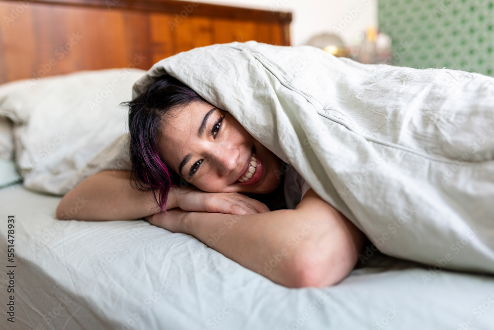 Happy young woman lying on bed with blanket at home Happy young woman lying on bed with blanket at home