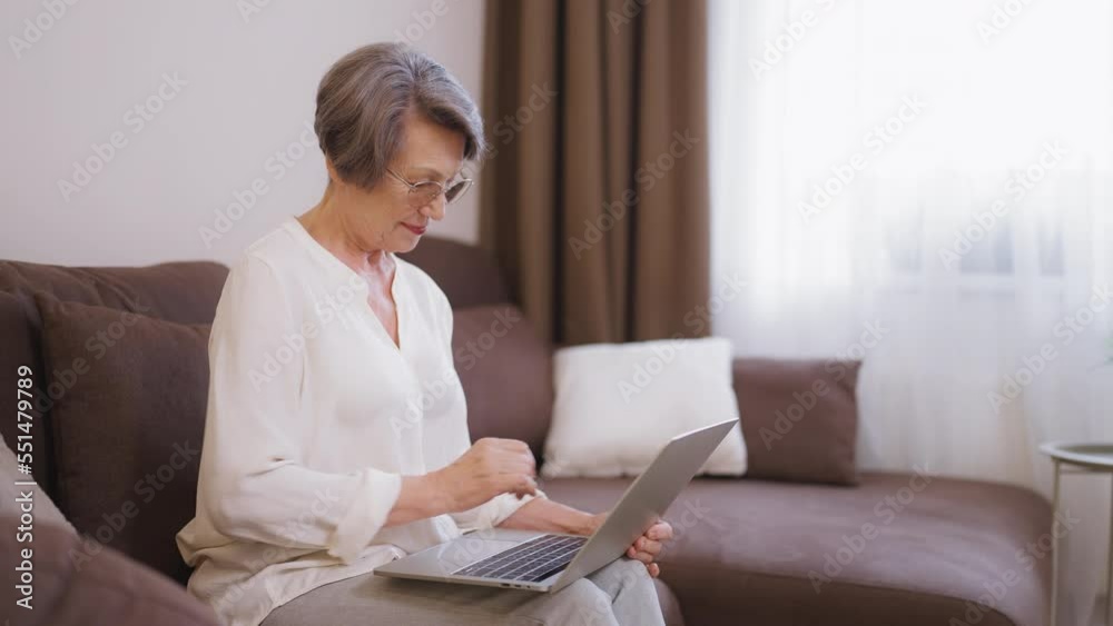 Senior lady in eyeglasses using laptop, sitting on couch at home, communication Senior lady in eyeglasses using laptop, sitting on couch at home, communication