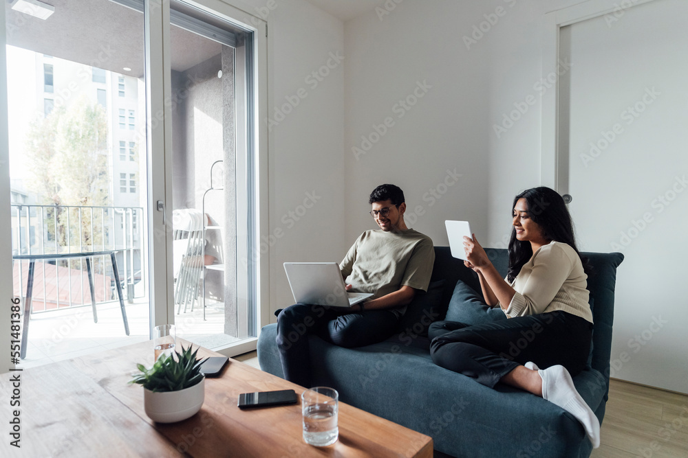 Young man and woman studying using laptop and tablet PC on sofa in living room Young man and woman studying using laptop and tablet PC on sofa in living room