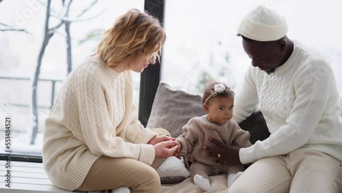 Happy interracial family sits on windowsill and plays with their baby daughter.