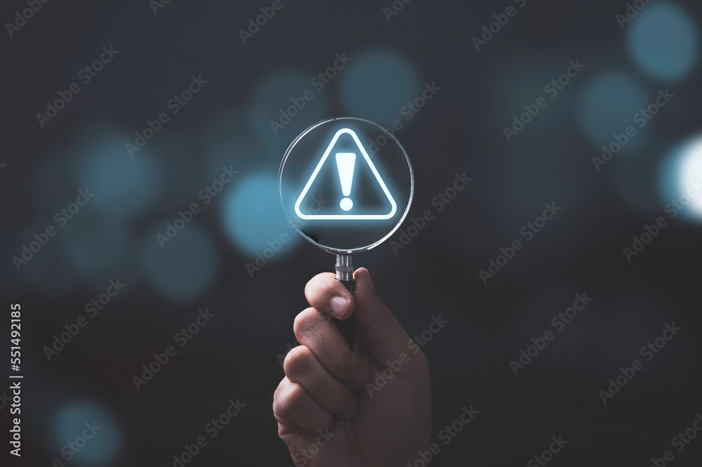 Hand holding magnifier glass with red triangle caution warning sign for notification error and maintenance concept. - obrazy, fototapety, plakaty 