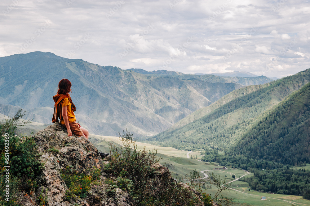 Side view of a red-haired girl sitting on a rock against the backdrop of a beautiful mountain valley