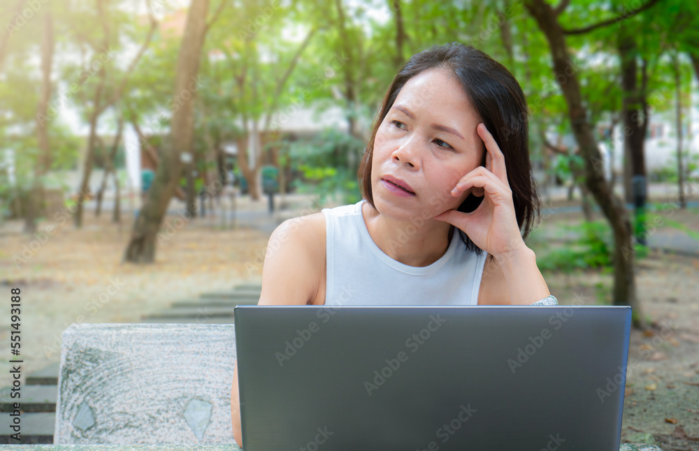 Asian middle-aged woman sitting at work and thinking or absent-minded at a desk with a laptop computer. Working in the garden, outdoor, wireless working concept. with technology Asian middle-aged woman sitting at work and thinking or absent-minded at a desk with a laptop computer. Working in the garden, outdoor, wireless working concept. with technology