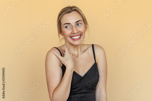 Shy, timid, pretty young woman with freckles stands over beige background. Beautiful girl surprised and wondered looks aside. Expressive facial expressions