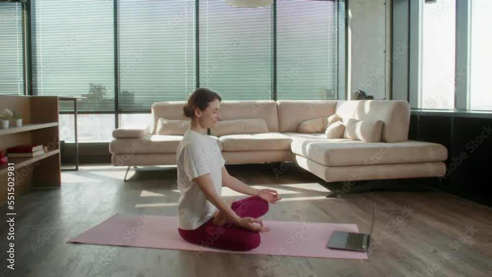 Middle age woman in sportswear practicing yoga at home. Female sit on mat in lotus position on red fitness mat in living room in front of open laptop, healthcare and exercise mental therapy at home