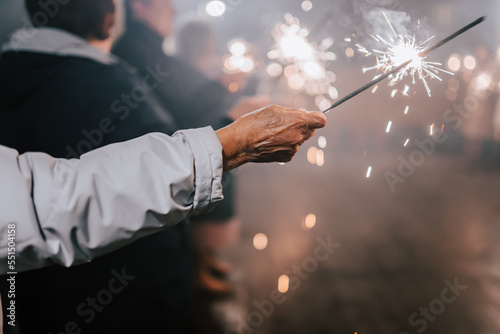 An old man holds a sparkling lights in his hand. Happy people with sparklers in their hands during celebration. Sparkler in hands on a wedding - guests holding lights in hand. 2023