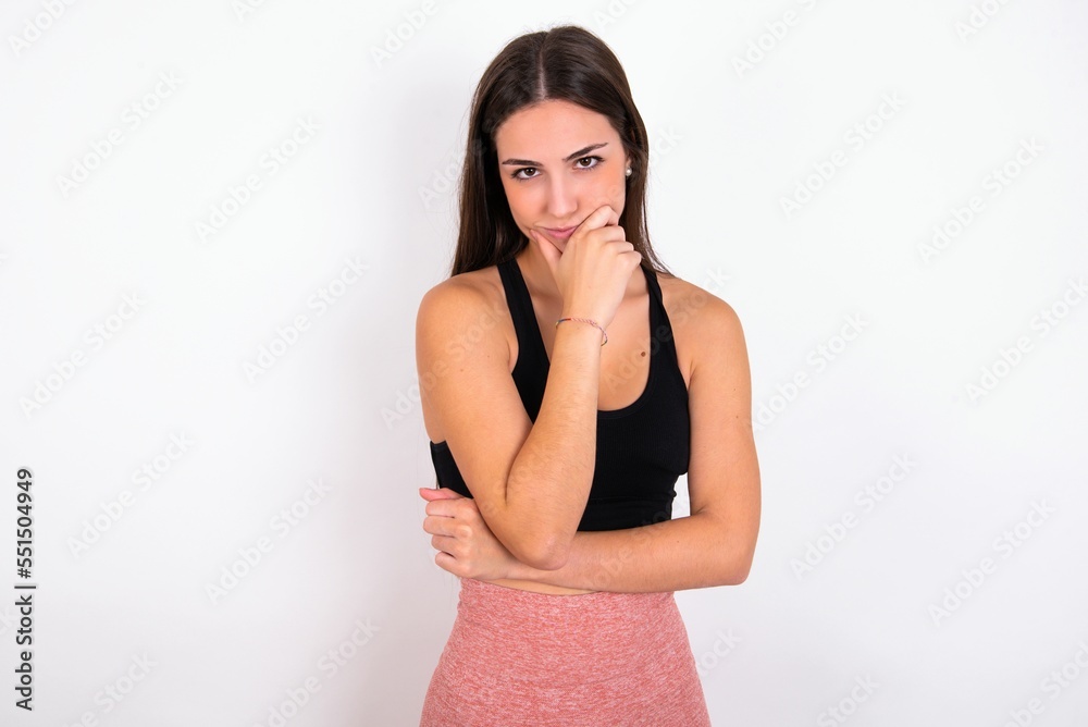 Thoughtful smiling Young caucasian woman wearing sportswear over white background keeps hand under chin, looks directly at camera, listens something with interest. Youth concept. Thoughtful smiling Young caucasian woman wearing sportswear over white background keeps hand under chin, looks directly at camera, listens something with interest. Youth concept.