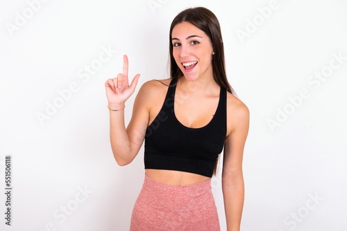 Pleasant looking Young caucasian woman wearing sportswear over white background  has clever expression, raises one finger, remembers himself not to forget tell important thing. © Jihan