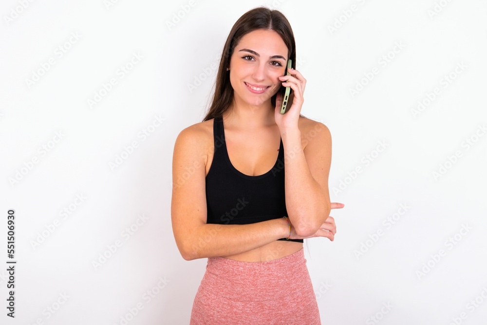 Portrait of a smiling Young caucasian woman wearing sportswear over white background talking on mobile phone. Business, confidence and communication concept. Portrait of a smiling Young caucasian woman wearing sportswear over white background talking on mobile phone. Business, confidence and communication concept.