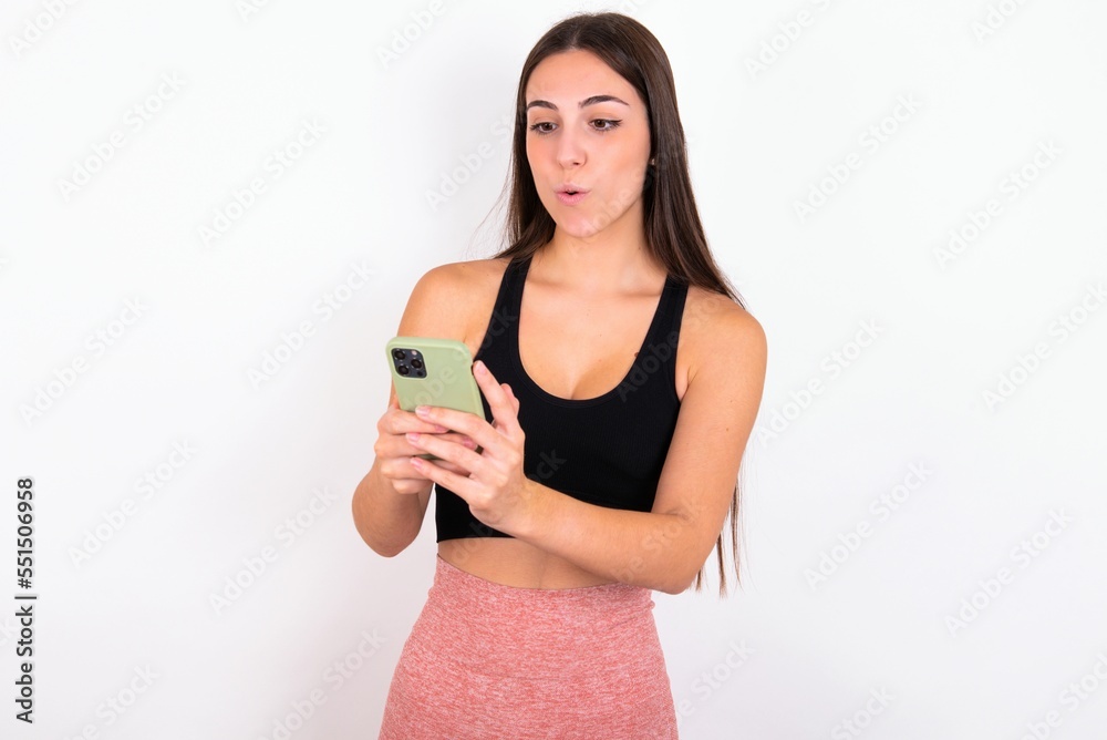 Young caucasian woman wearing sportswear over white background looks with bugged eyes, holds modern smart phone, receives unexpected message from friend, reads reminder. Young caucasian woman wearing sportswear over white background looks with bugged eyes, holds modern smart phone, receives unexpected message from friend, reads reminder.