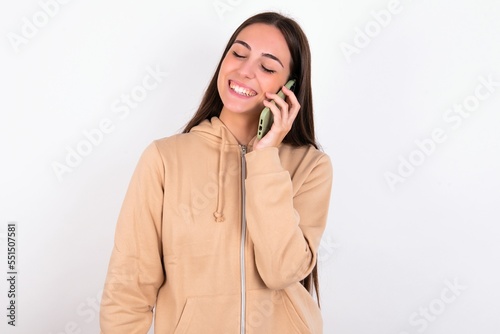 Funny Young caucasian woman wearing sweatshirt over white background laughs happily, has phone conversation, being amused by friend, closes eyes.