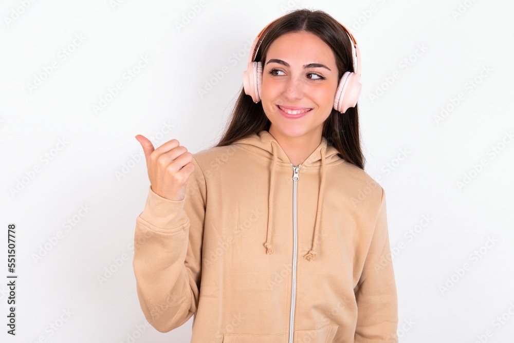 Young caucasian woman wearing sweatshirt over white background listens audio track via wireless headphones points thumb away advertises copy space Young caucasian woman wearing sweatshirt over white background listens audio track via wireless headphones points thumb away advertises copy space