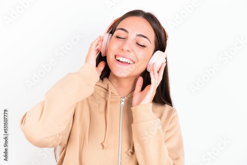 Young caucasian woman wearing sweatshirt over white background smiles broadly feels very glad listens favourite music track via wireless headphones closes eyes.