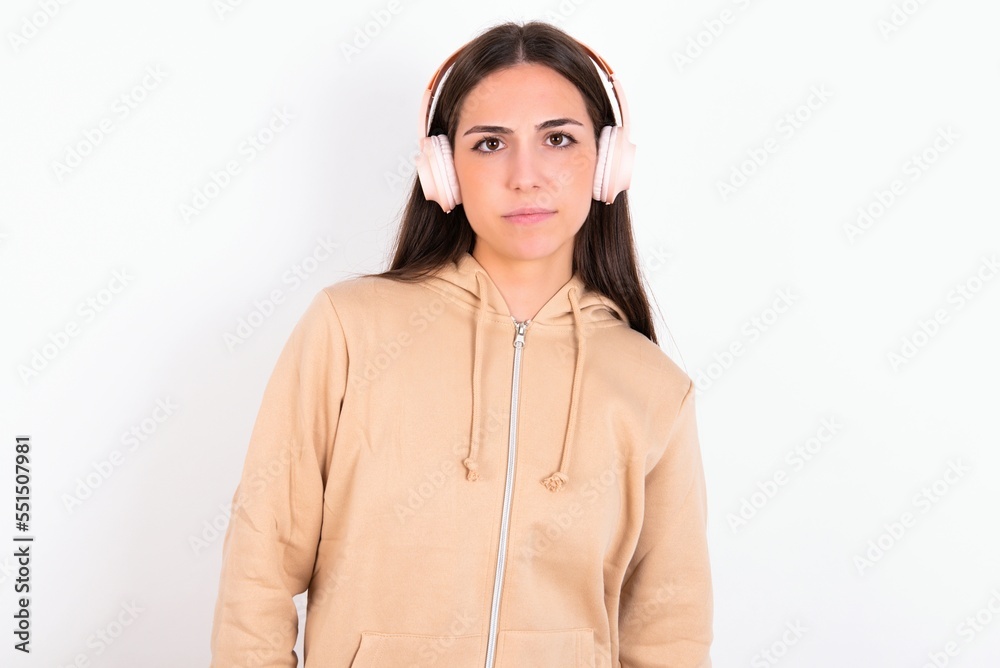 Serious displeased Young caucasian woman wearing sweatshirt over white background looks puzzled at camera being angry wears stereo headphones listens music while walking at street Serious displeased Young caucasian woman wearing sweatshirt over white background looks puzzled at camera being angry wears stereo headphones listens music while walking at street