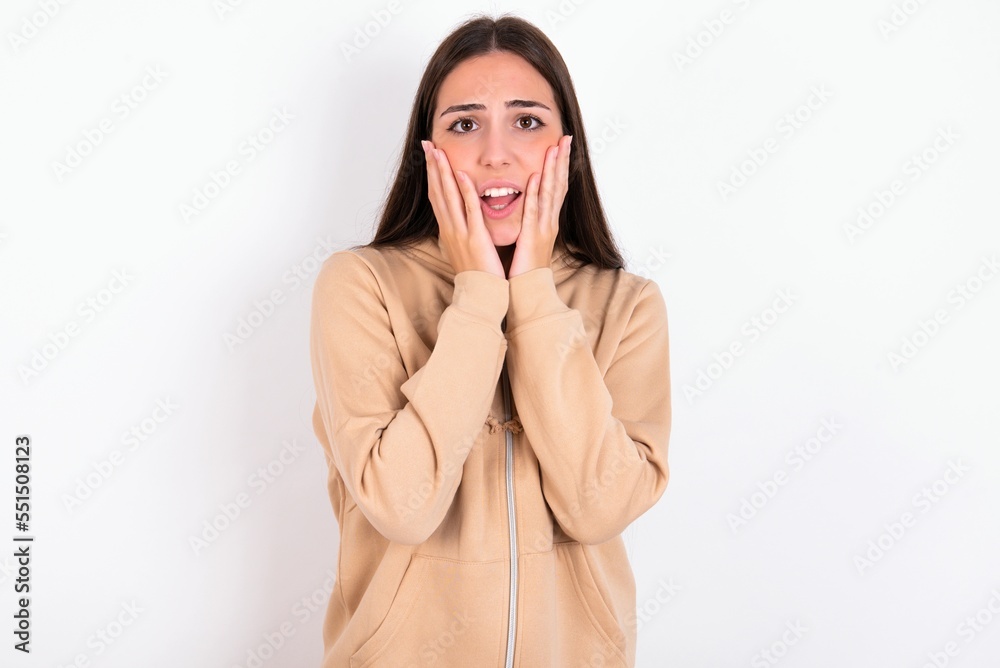Upset Young caucasian woman wearing sweatshirt over white background touching face with two hands Upset Young caucasian woman wearing sweatshirt over white background touching face with two hands