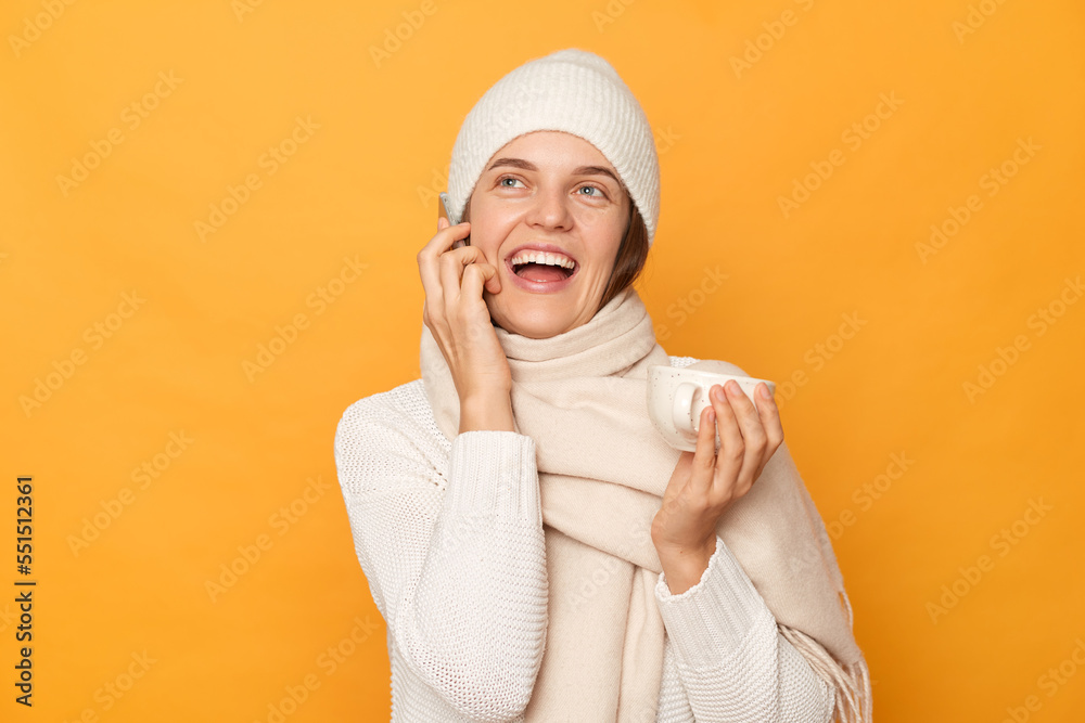 Indoor shot of joyful beautiful Caucasian woman wearing warm jumper, cap and scarf, talking on cell phone with happy expression, drinking coffee, posing isolated over yellow background. Indoor shot of joyful beautiful Caucasian woman wearing warm jumper, cap and scarf, talking on cell phone with happy expression, drinking coffee, posing isolated over yellow background.