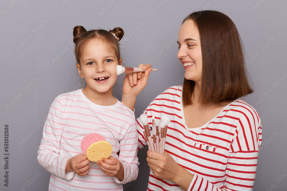 Horizontal shot of smiling young adult brown hair woman with her daughter posing isolated over gray background, mother applying powder with brush to her kid's cheek. Horizontal shot of smiling young adult brown hair woman with her daughter posing isolated over gray background, mother applying powder with brush to her kid's cheek.