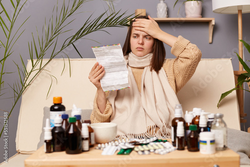 Image of unhealthy ill Caucasian woman reading instructions before taking medication, suffering terrible headache, touched her forehead, having high temperature.