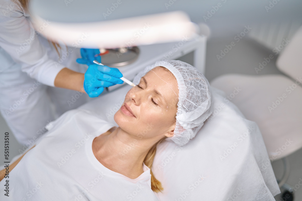 Cosmetologist drawing mark on client face to prepare for rejuvenation treatment Cosmetologist drawing mark on client face to prepare for rejuvenation treatment