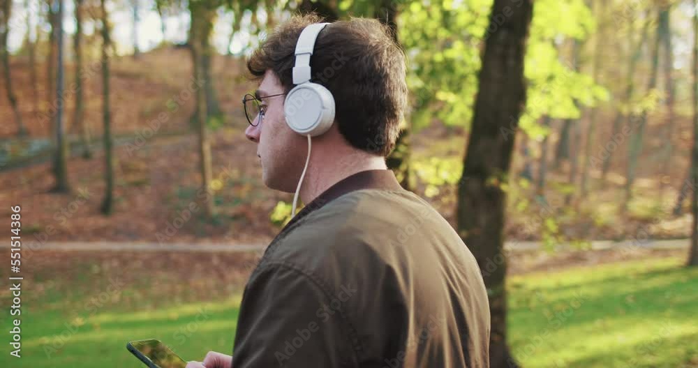 Caucasian man in headphones and glasses walking in park among trees in autumn, listening to music and using smartphone. Young male in eyeglasses strolling and holding cellphone. Caucasian man in headphones and glasses walking in park among trees in autumn, listening to music and using smartphone. Young male in eyeglasses strolling and holding cellphone.