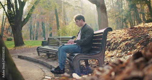 Caucasian young man sitting on bench in park, opening laptop and start working. Outdoor. Male freelancer typing on computer in nature in fall. Resting and chatting online. Remote work concept. photo